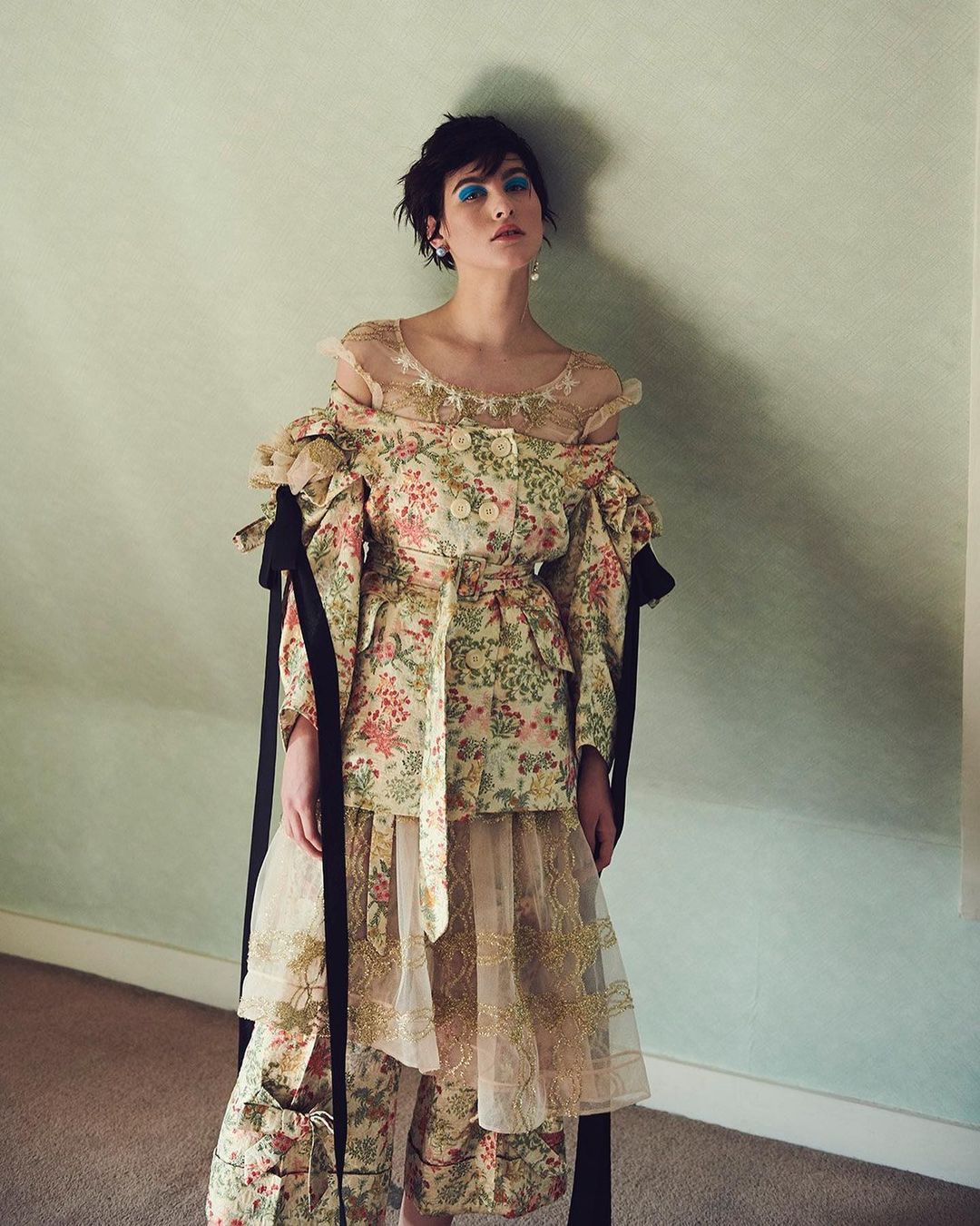A woman with short dark hair wearing a floral patterned outfit, poses indoors against a light green wall in a medium shot.