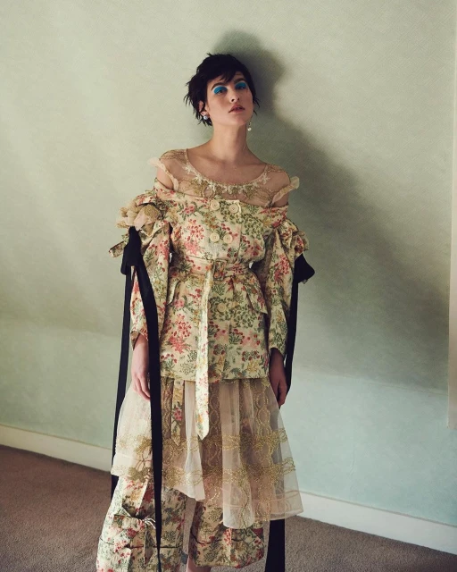 A woman with short dark hair wearing a floral patterned outfit, poses indoors against a light green wall in a medium shot.