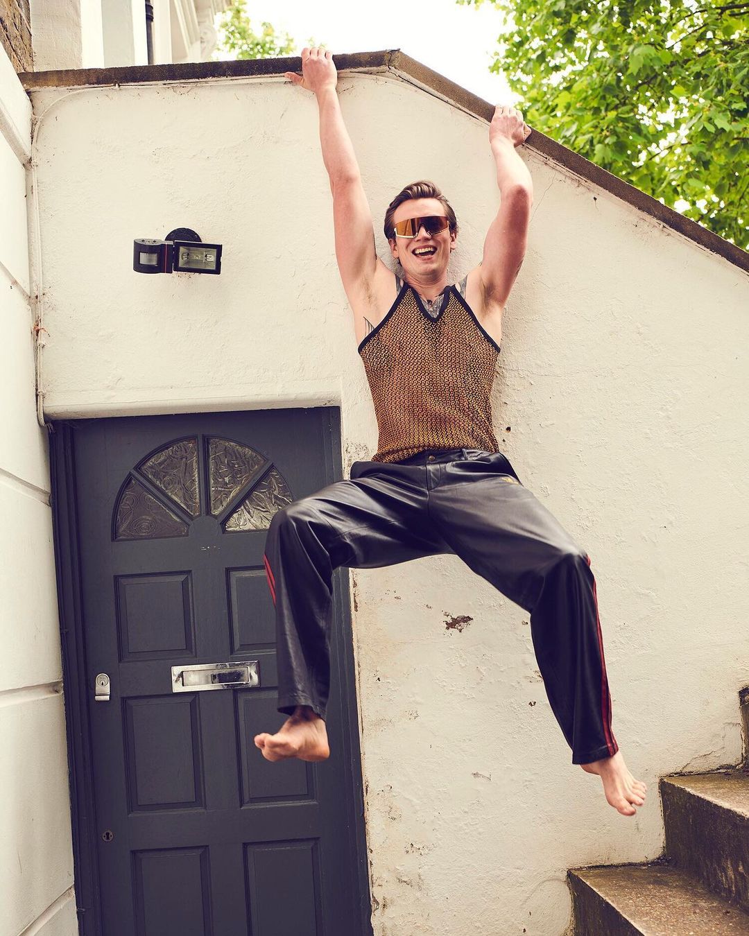 Athletic man in sunglasses and mesh top hanging from ledge outdoors, wearing sweatpants, barefoot, with a door and steps in the background.