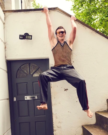 Athletic man in sunglasses and mesh top hanging from ledge outdoors, wearing sweatpants, barefoot, with a door and steps in the background.