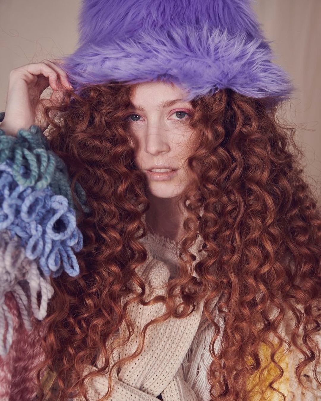 Close-up portrait of a fair-skinned woman with voluminous red curly hair wearing a lavender faux fur hat and a knitted sweater.