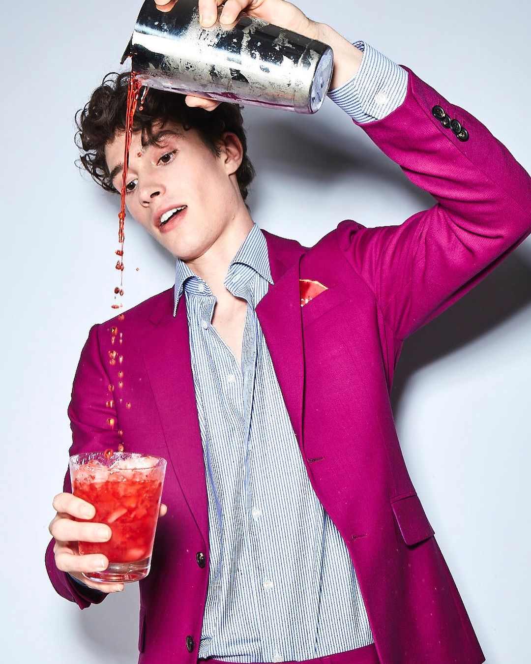 Man in magenta suit prepares a cocktail. He's pouring a red drink from a silver cocktail shaker into a glass filled with ice.