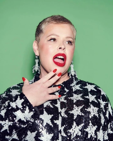 Close-up portrait of a woman with short blonde hair wearing a sequined star-patterned top, red lipstick, and ornate dangling earrings
