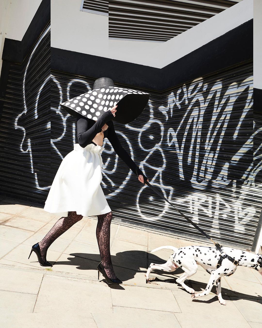Woman in polka dot hat walks a Dalmatian in an urban setting. She wears a white skirt, patterned stockings, and black heels. Graffiti background.