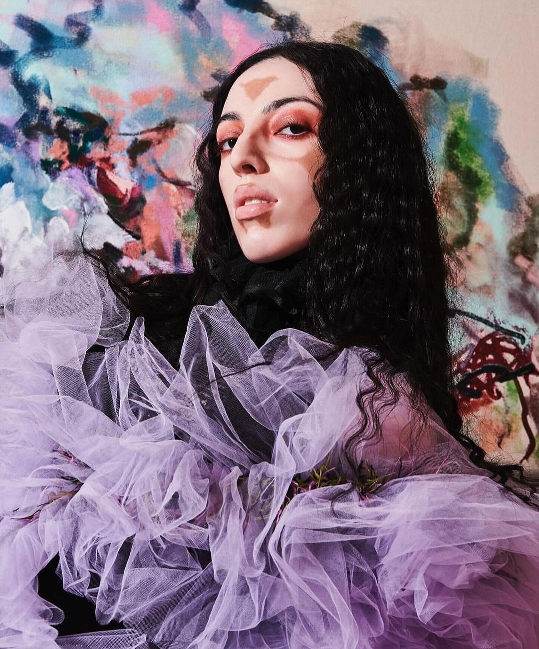 Woman poses in a violet tulle dress with colorful face paint and expressive makeup, set against an abstract artistic background.