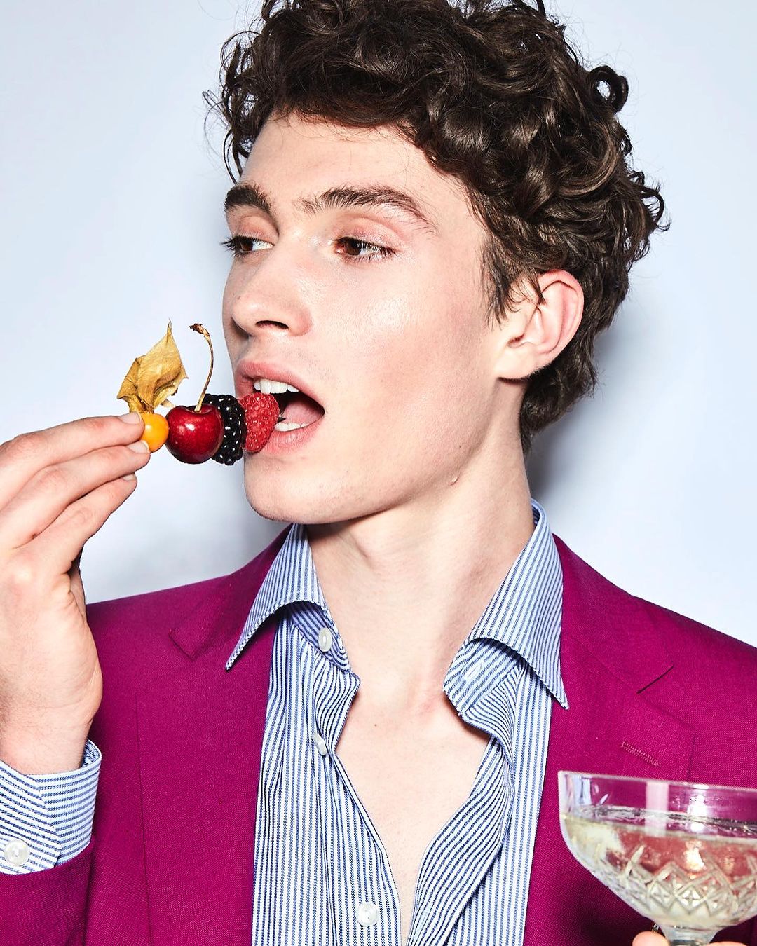 Handsome man in a fuchsia suit holds berries on a stick near his mouth, and a champagne glass, in a close-up, indoors.