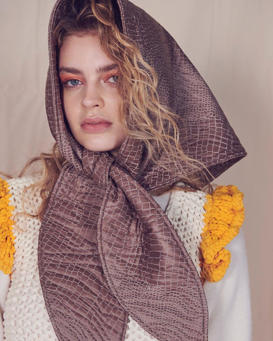 Woman wearing an auburn headscarf with a crocodile print and a white sweater with yellow pom poms posing in a close-up shot.