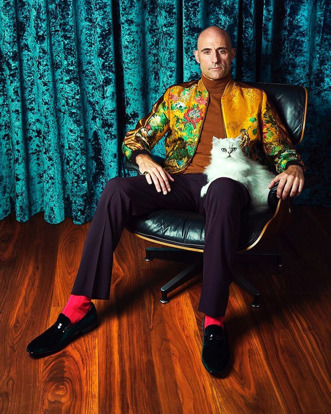 Portrait of a stylish adult male in a floral jacket sitting in a black chair with a white cat in his lap in an indoor setting.