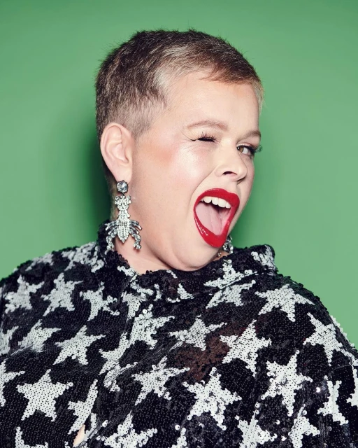 Close-up portrait of a winking woman with short blonde hair, red lipstick, and beetle earrings against a solid green backdrop.