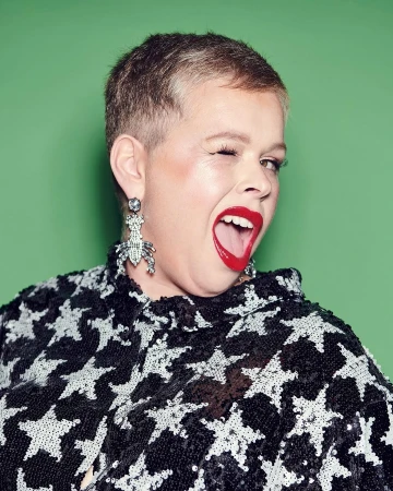 Close-up portrait of a winking woman with short blonde hair, red lipstick, and beetle earrings against a solid green backdrop.