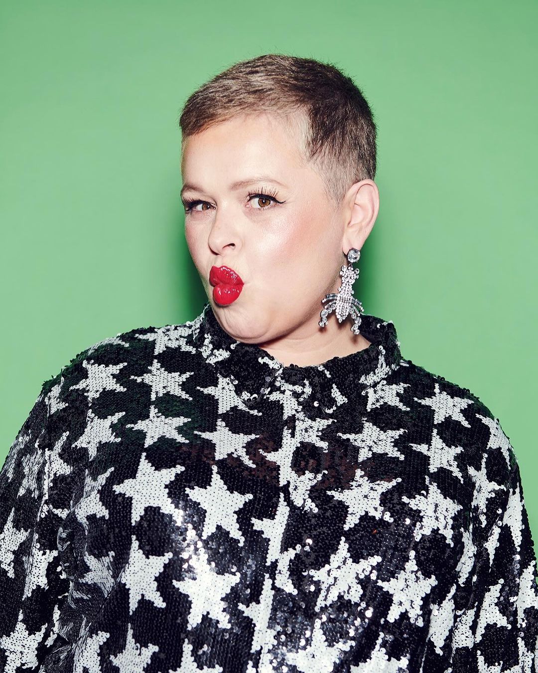 A woman with short brown hair wearing a black sequined star-print top and statement earrings puckers her red lips against a green background.