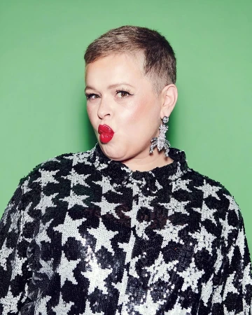 A woman with short brown hair wearing a black sequined star-print top and statement earrings puckers her red lips against a green background.