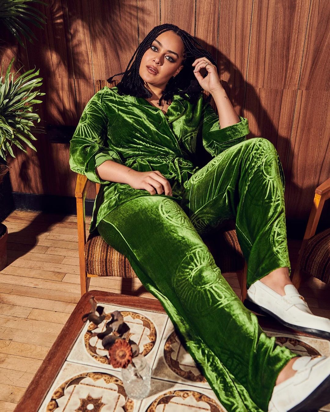 Woman in a green velvet patterned pantsuit sits indoors, hand near head, wearing white loafers, beside a patterned tile table.