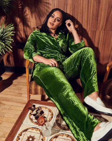 Woman in a green velvet patterned pantsuit sits indoors, hand near head, wearing white loafers, beside a patterned tile table.