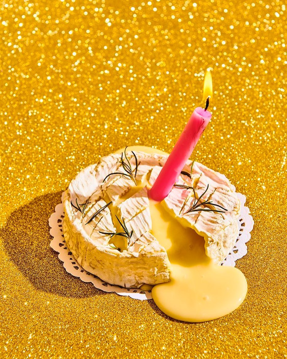 Novel dessert presentation: Brie cheese with melted center, topped with herbs and a lit candle, set against a sparkly gold background.