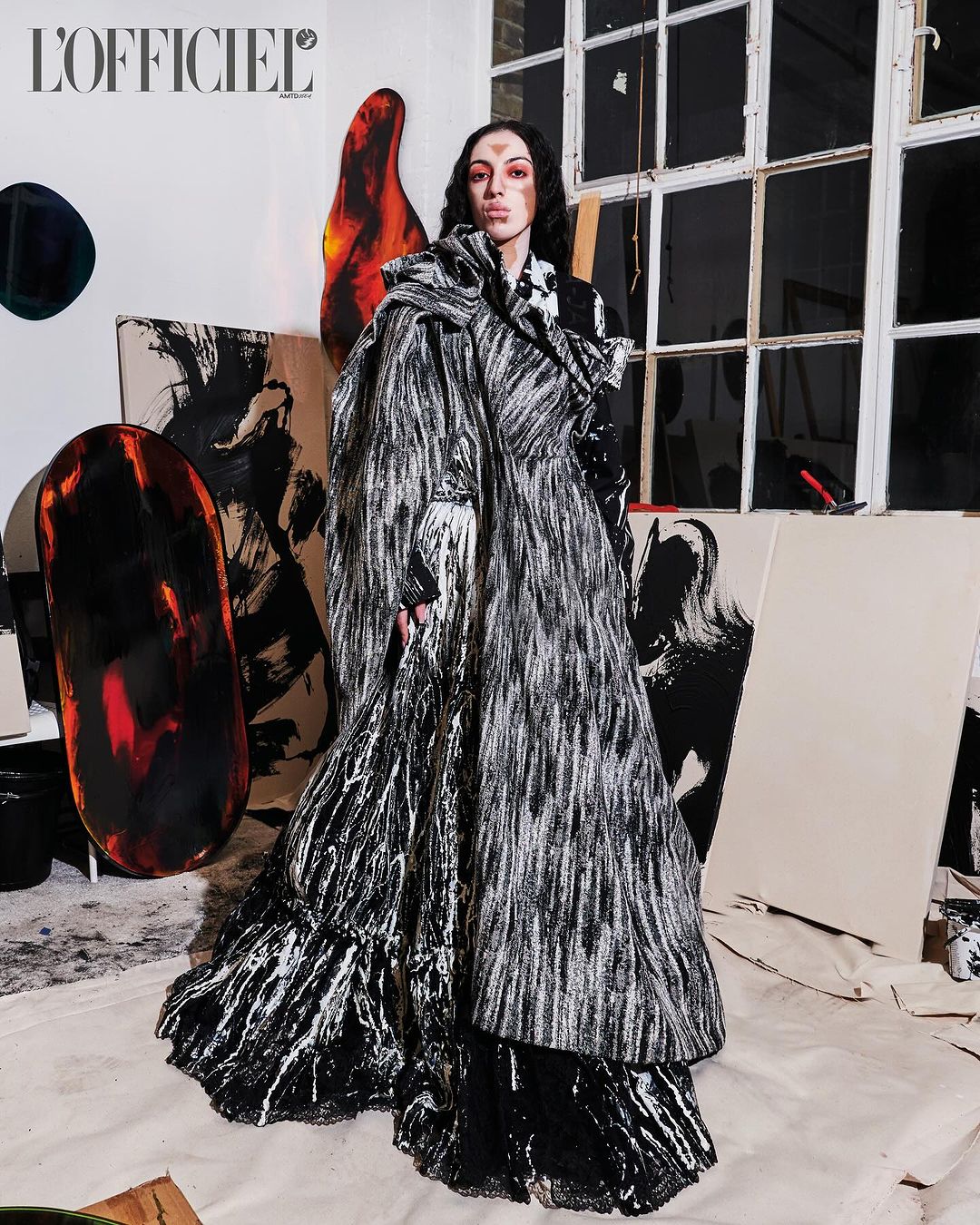 A woman poses in a long black and white coat with abstract paintings in the background, indoor, studio, fashion editorial photo.