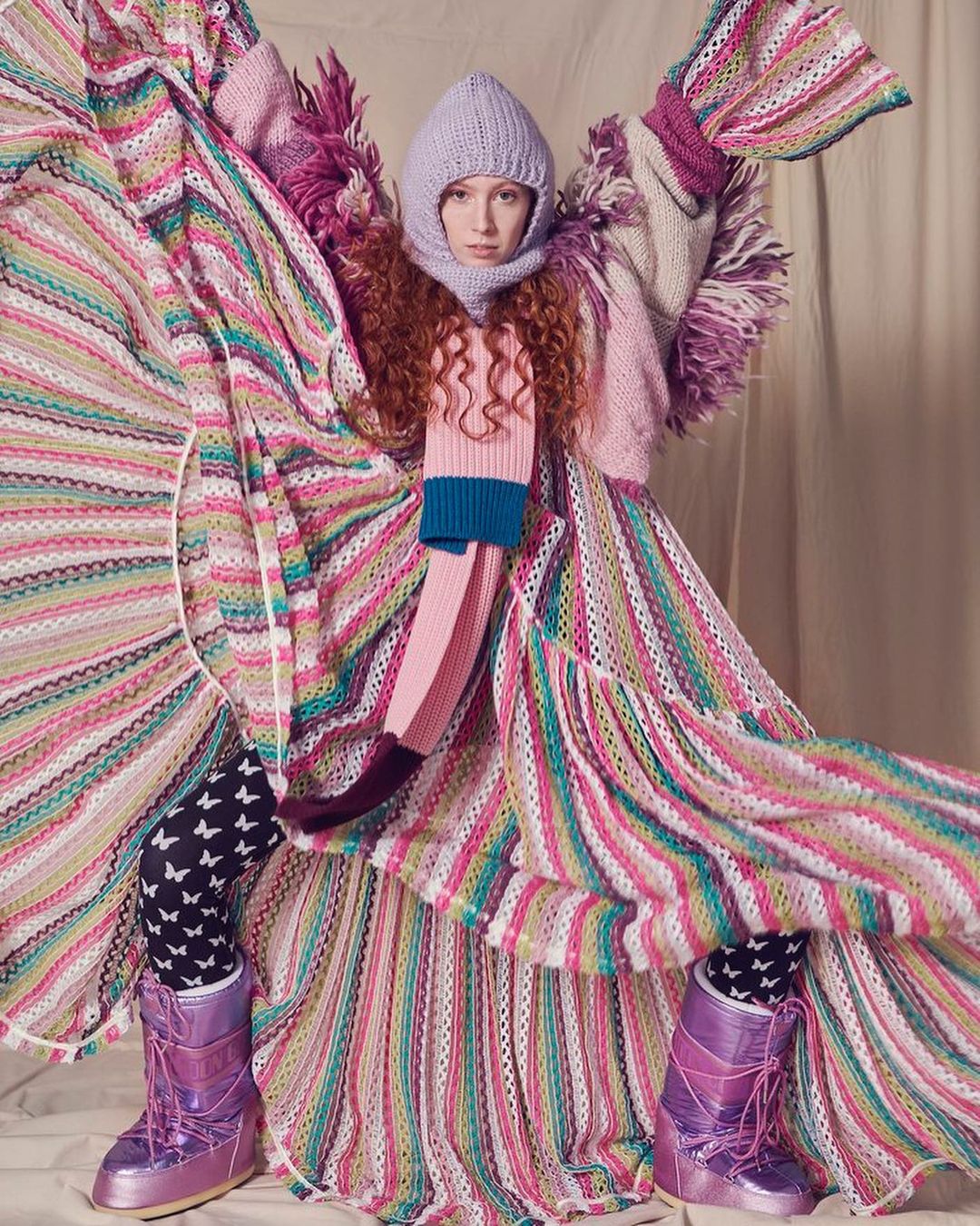 Fashion shot of woman in colorful knitwear including a long dress, scarf, and balaclava, paired with iridescent purple moon boots.