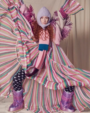 Fashion shot of woman in colorful knitwear including a long dress, scarf, and balaclava, paired with iridescent purple moon boots.