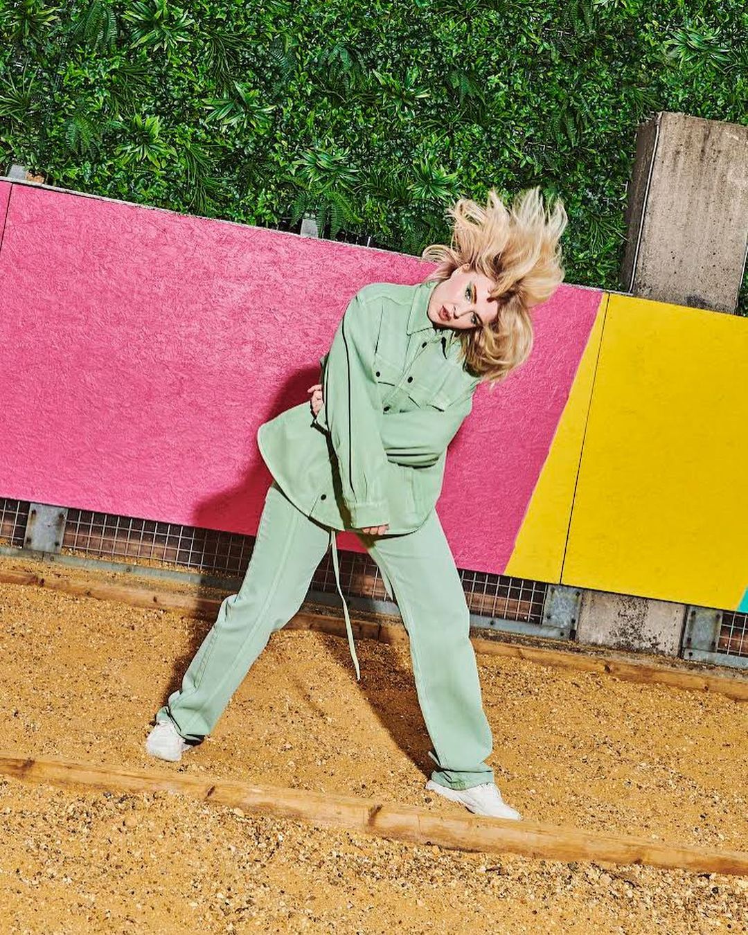 Young woman with blonde hair poses in a light green suit with colorful geometric shapes and green foliage in the background outdoors.