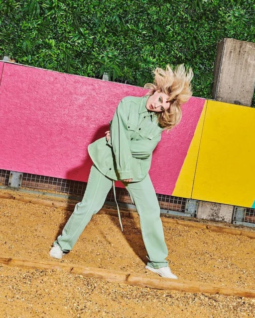 Young woman with blonde hair poses in a light green suit with colorful geometric shapes and green foliage in the background outdoors.