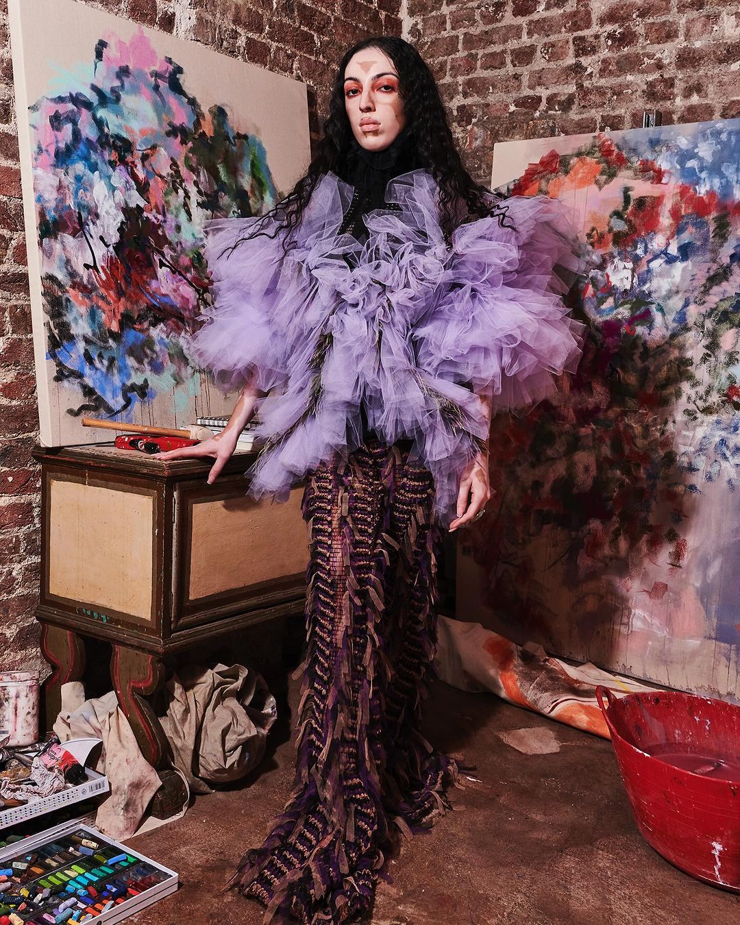 A woman in a lilac tulle top and brown fringe dress poses near paintings and art supplies, showcasing avant-garde fashion in an artist studio.