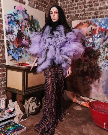 A woman in a lilac tulle top and brown fringe dress poses near paintings and art supplies, showcasing avant-garde fashion in an artist studio.