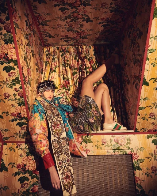 Two women pose in a room covered in floral wallpaper. One woman sits with legs raised, showcasing designer shoes. The other wears a patterned coat.
