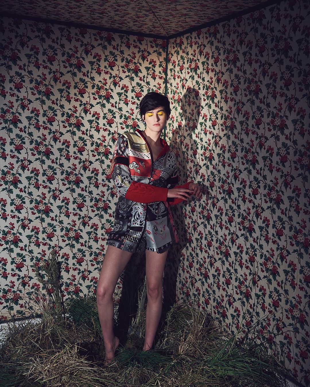 Woman poses in an artistic studio portrait, wearing avant-garde fashion, with vivid makeup and a striking floral backdrop and organic ground cover.