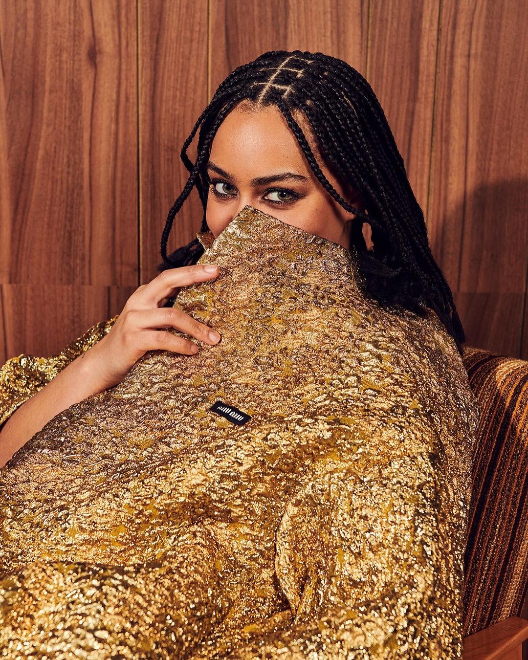 Portrait of a woman with braided hair wearing a textured gold jacket, partially obscuring her face, against a wood panel background.