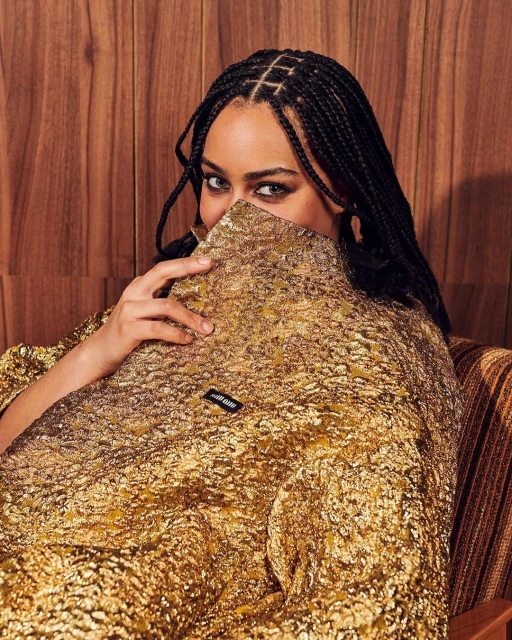 Portrait of a woman with braided hair wearing a textured gold jacket, partially obscuring her face, against a wood panel background.