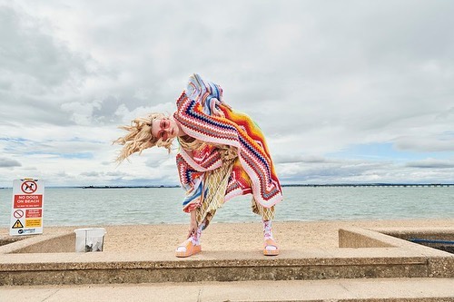 Woman in colorful knitted blanket and sandals poses on a concrete platform in front of a beach, with a cloudy sky in the background.