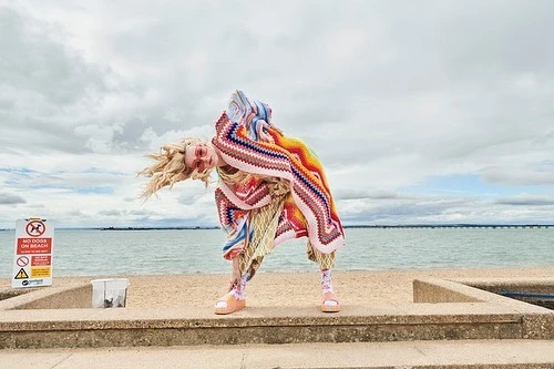 Woman in colorful knitted blanket and sandals poses on a concrete platform in front of a beach, with a cloudy sky in the background.