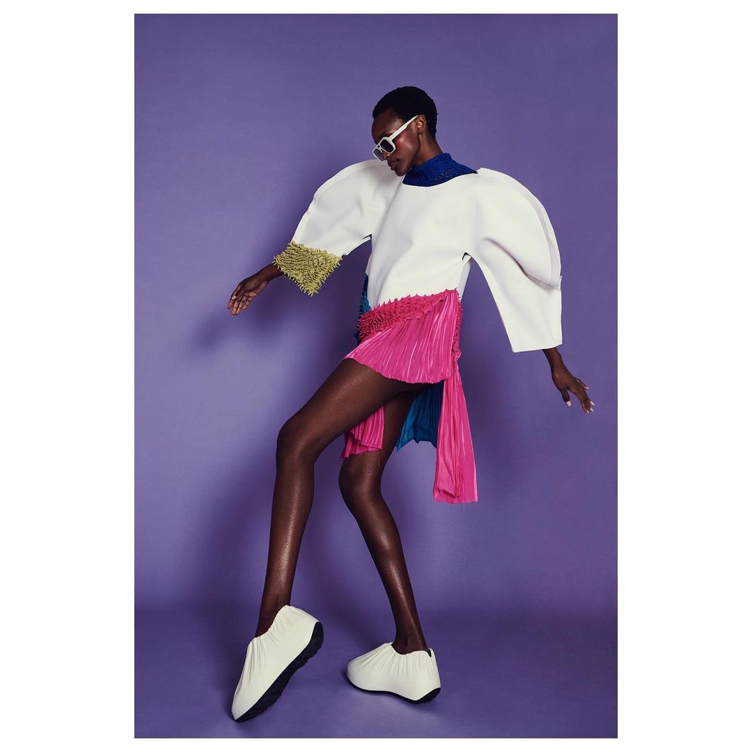 A slender African American woman strikes a fashion pose in a studio, wearing a white top, pink skirt, white shoes, and sunglasses against a purple backdrop.