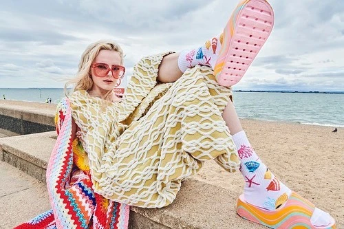 A blonde woman in a patterned yellow outfit and colorful cardigan poses on a beachside wall with ocean and sky in the background.