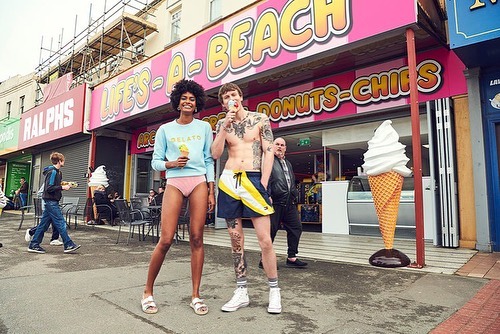 A shirtless man and an African American woman pose with ice cream cones on a sidewalk in front of a beachside shop on a sunny day.