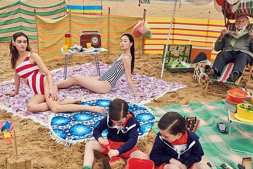Group of people on a sandy beach with striped windbreaks, featuring adult women in swimsuits and children playing in the sand.
