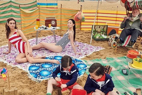 Group of people on a sandy beach with striped windbreaks, featuring adult women in swimsuits and children playing in the sand.