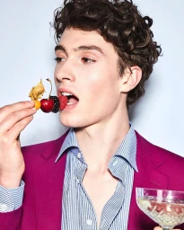 Young man with curly brown hair wearing a pink blazer eating a skewer of fresh berries against a white background