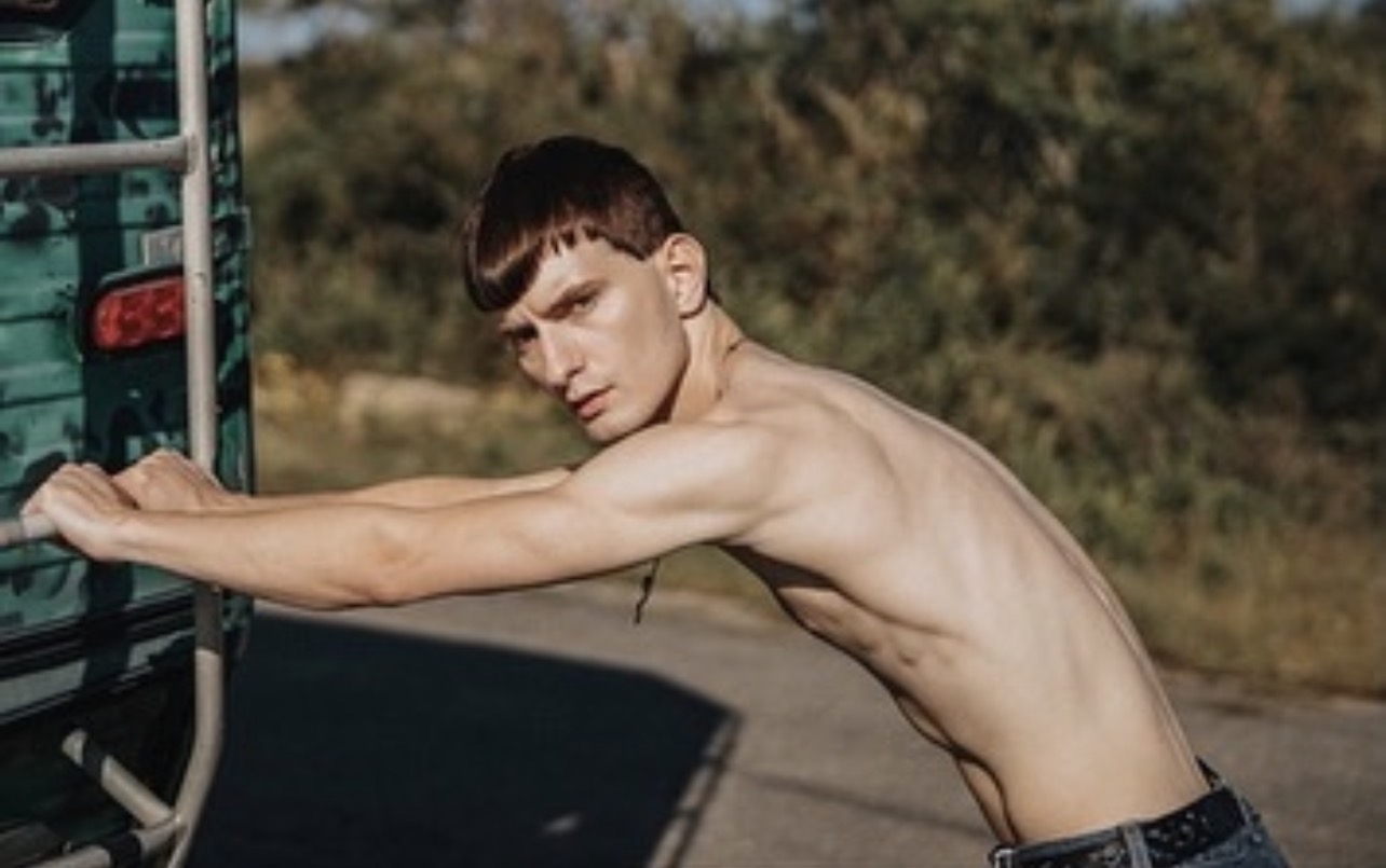 Shirtless male model with cropped dark hair leaning forward against the metal ladder of a vintage teal van on an open road.