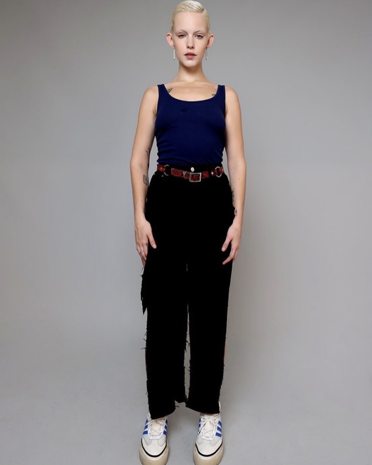 Model with blonde hair wearing a blue tank top, black pants, and a patterned belt against a plain grey background
