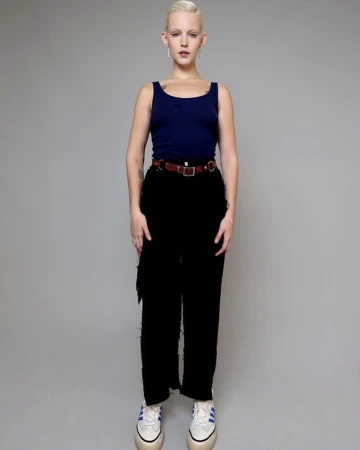 Model with blonde hair wearing a blue tank top, black pants, and a patterned belt against a plain grey background