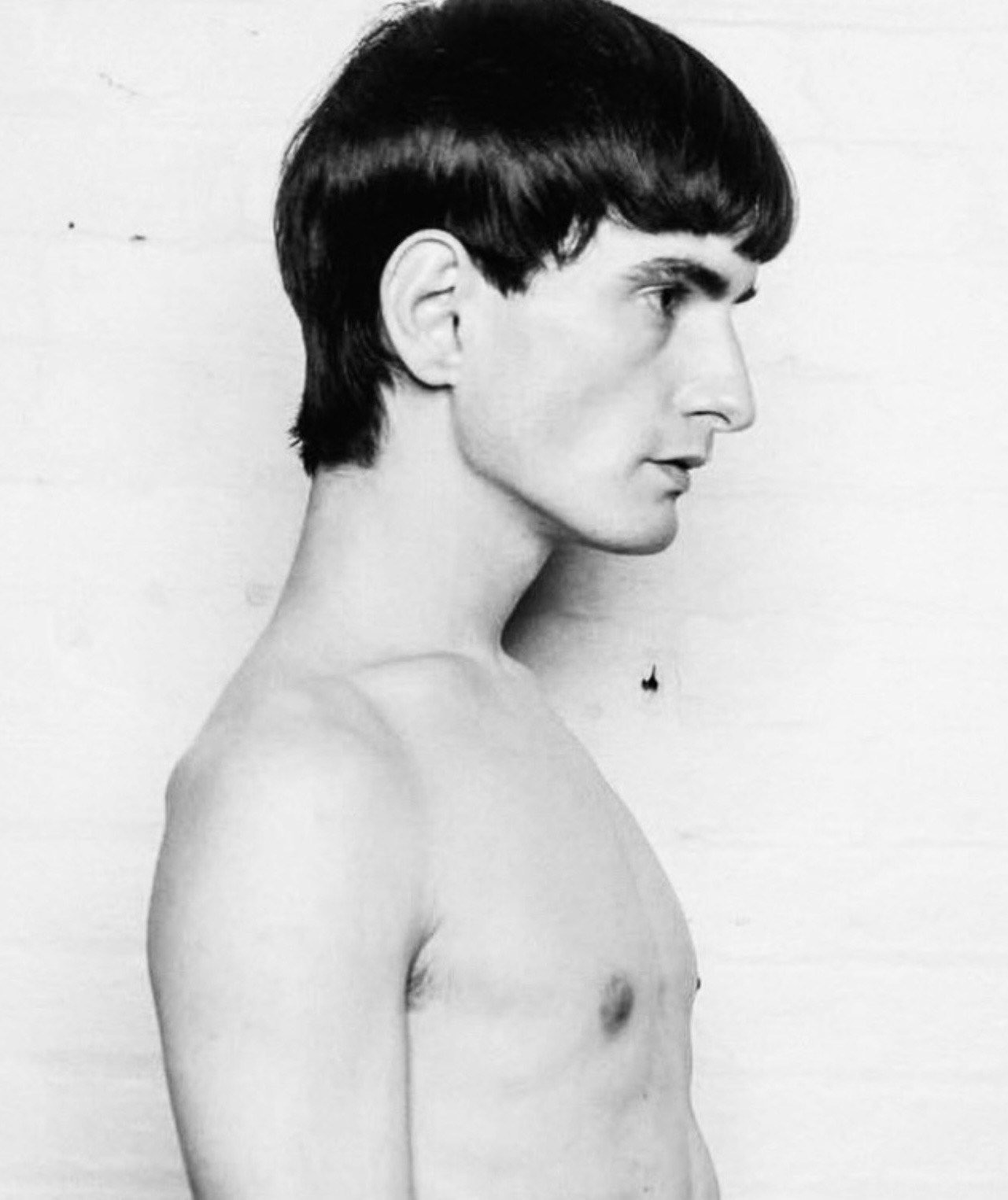 Black and white profile portrait of a young male model with dark bowl cut hair against a plain studio background.