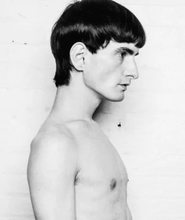 Black and white profile portrait of a young male model with dark bowl cut hair against a plain studio background.