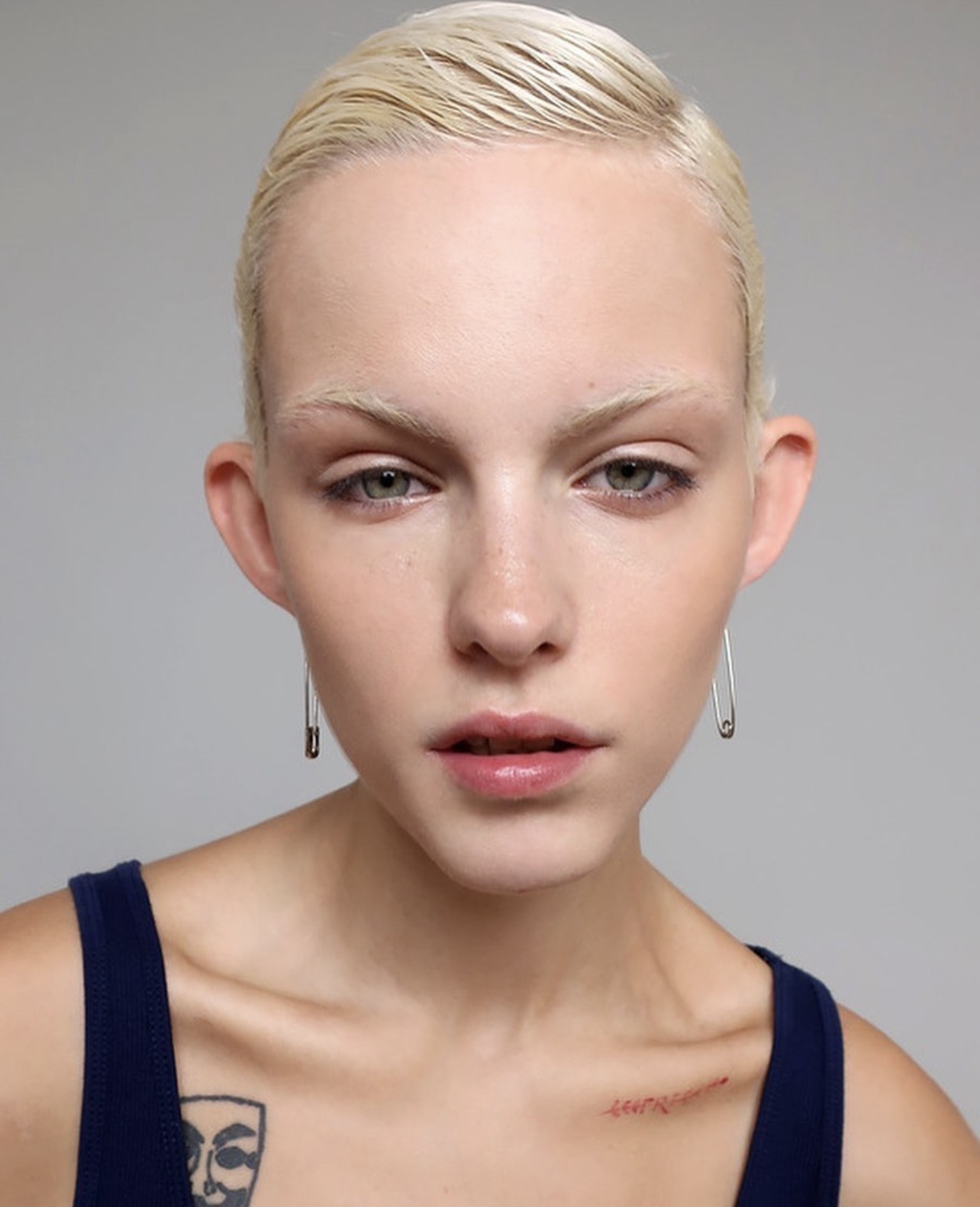 Close-up portrait of a young adult female with bleached blonde hair and fair skin, wearing safety-pin earrings against a neutral backdrop.