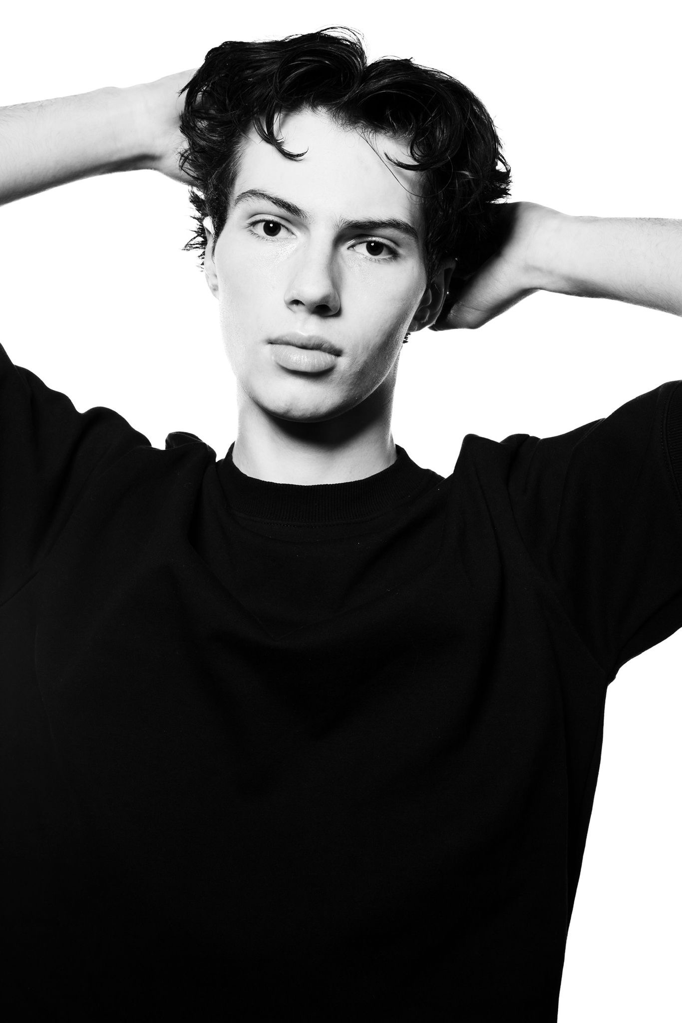 Portrait of a young man with dark curly hair posing with hands behind his head, wearing a black t-shirt against a bright white background.
