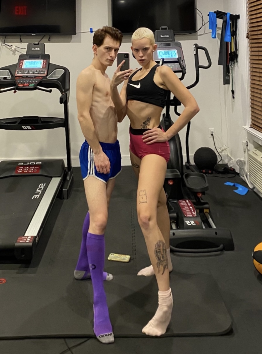 Two athletic people posing for a cell phone photo in a home gym setting with treadmills and exercise equipment in the background.