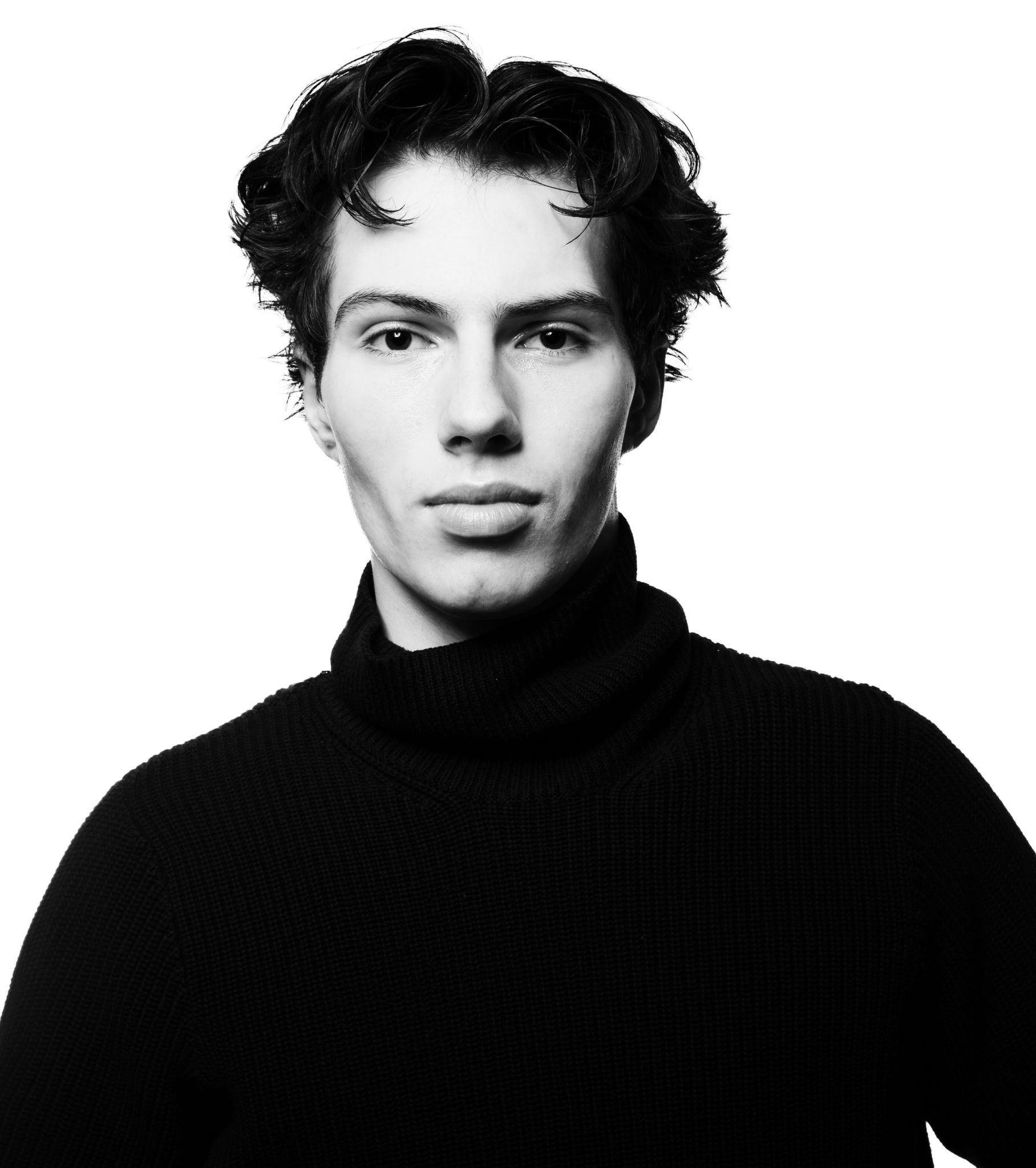 Black and white portrait of a young adult male with dark curly hair, wearing a black turtleneck sweater, against a white background.