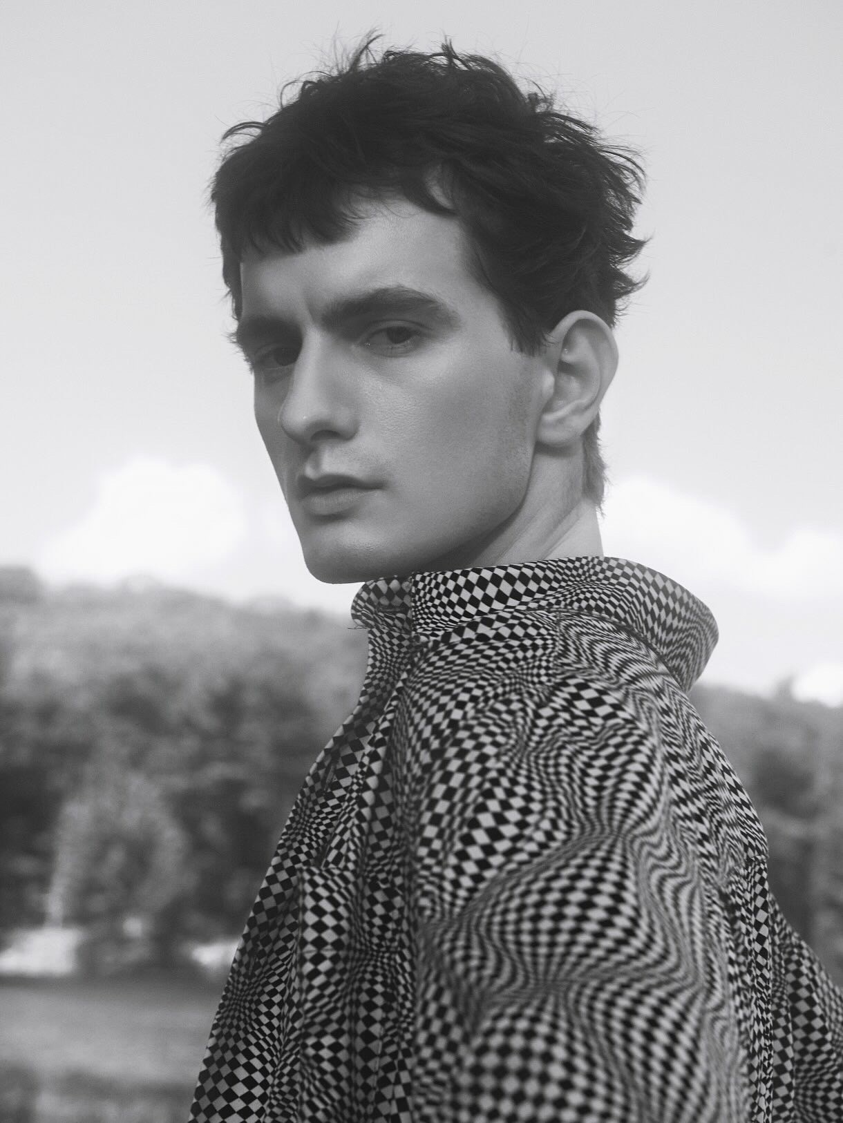 Monochrome portrait of a young man with dark tousled hair wearing a patterned checkered high-collar shirt outdoors.