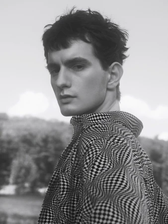 Monochrome portrait of a young man with dark tousled hair wearing a patterned checkered high-collar shirt outdoors.