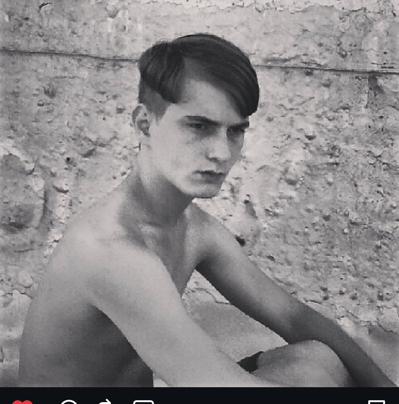 Black and white portrait of a young adult male with a side swept haircut, seated outdoors against a textured stone wall in natural light.